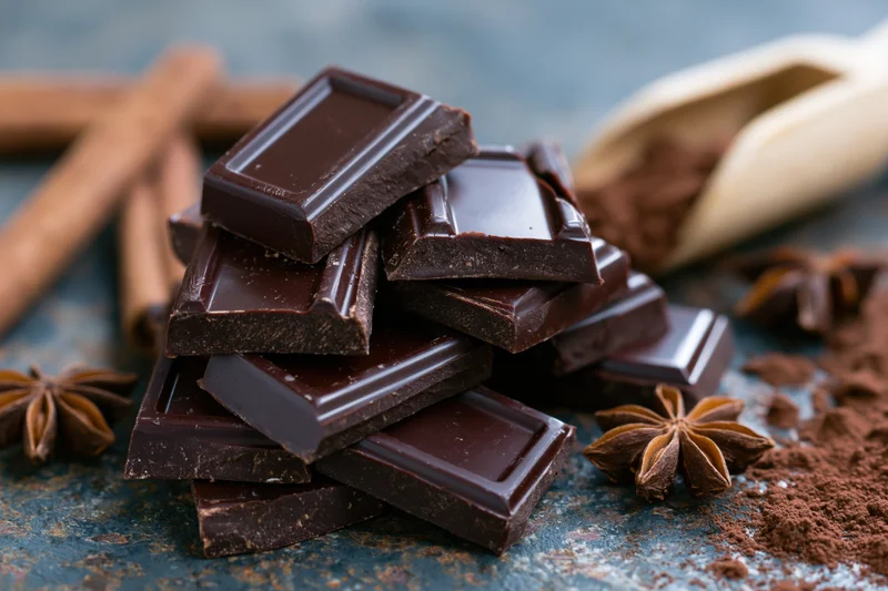 Broken dark chocolate bar pieces stacked on a rustic blue surface with cinnamon sticks, star anise, and cocoa powder