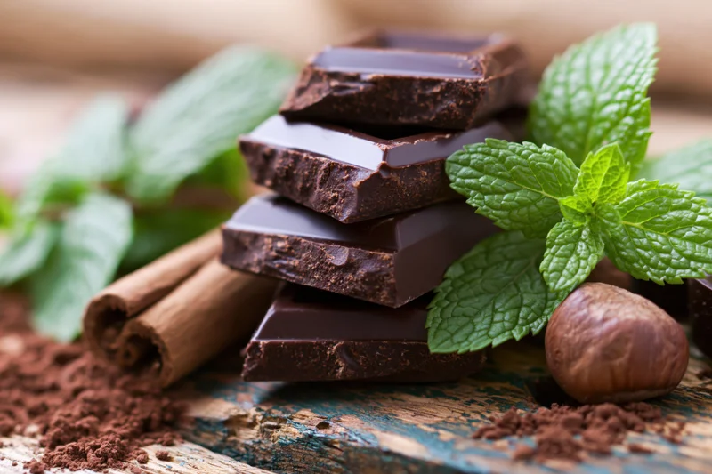 Stacked dark chocolate pieces with fresh mint, cinnamon sticks, and hazelnuts on a rustic wooden surface
