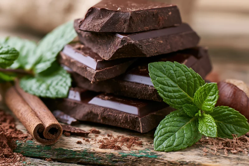 Stacked dark chocolate pieces with fresh mint and cinnamon sticks on a rustic wooden surface