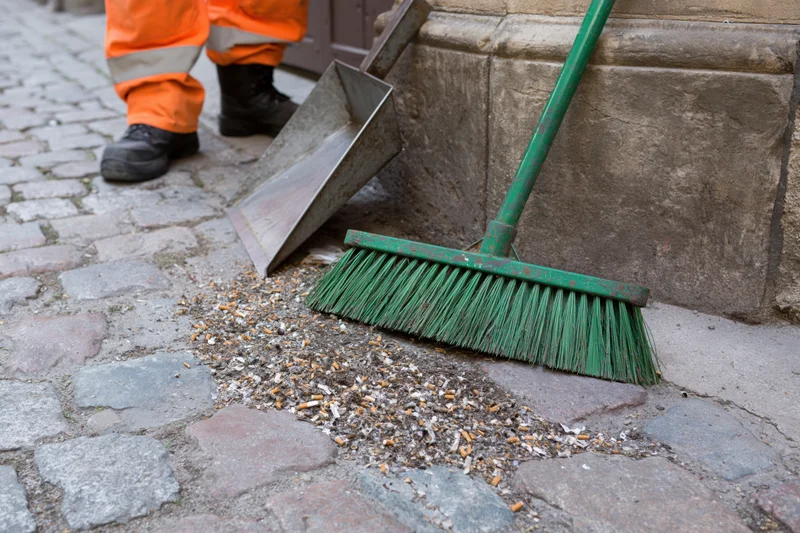 Street cleaner sweeping cigarette butts and small litter into a dustpan on a cobblestone sidewalk
