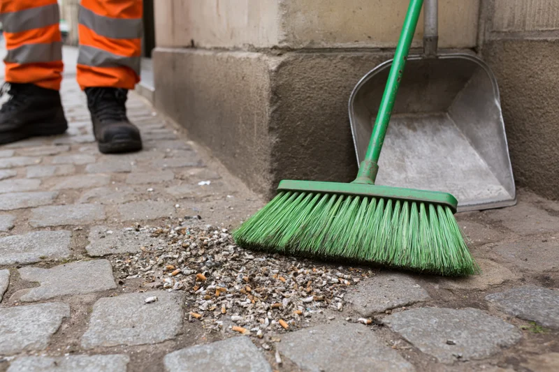 Street cleaner sweeping cigarette butts and small litter into a dustpan on a cobblestone sidewalk