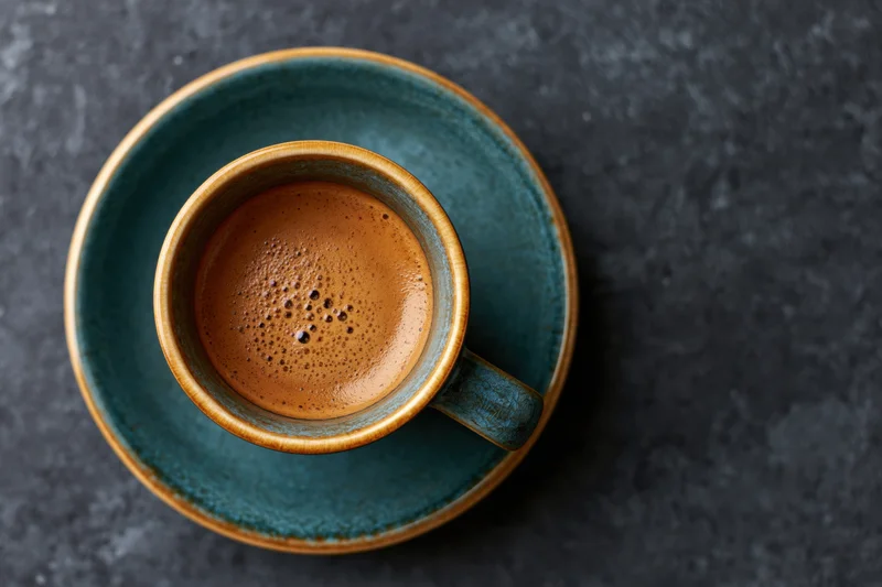 Overhead view of a teal ceramic espresso cup with foamy crema on a dark textured surface