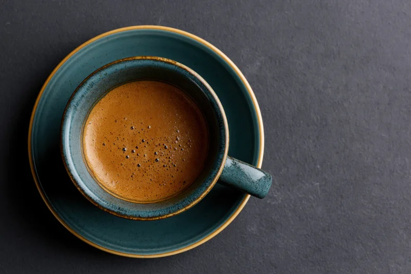 Teal ceramic cup of espresso with foamy crema on dark surface, overhead view