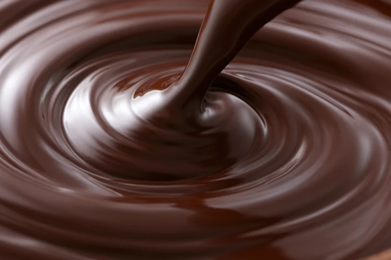 Thick stream of melted dark chocolate pouring into a swirling pool, creating ripples and glossy reflections
