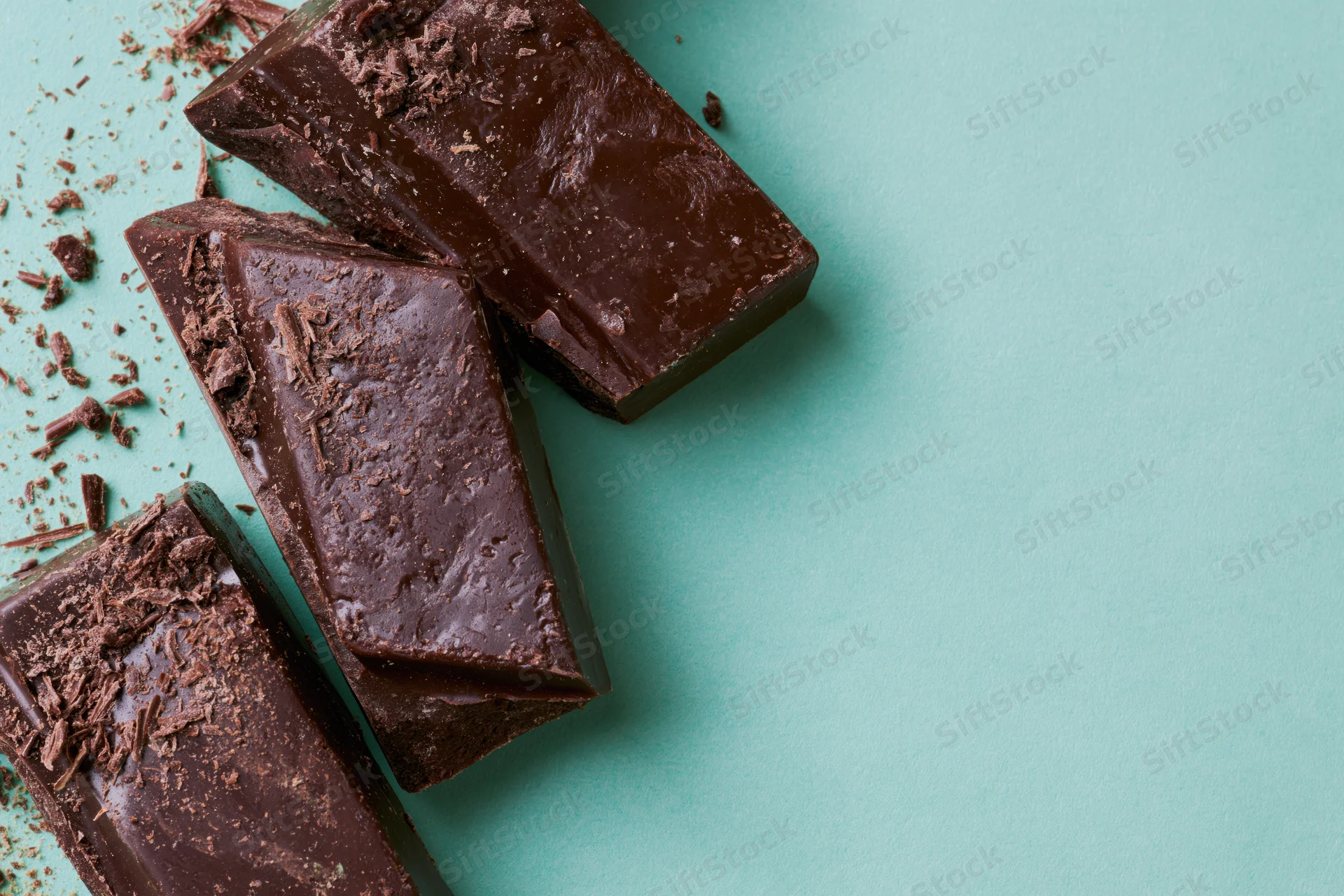 Broken dark chocolate chunks arranged vertically on a teal surface with scattered shavings and copyspace