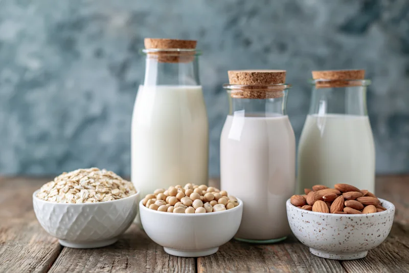 Three glass bottles of plant-based milk with bowls of oats, soybeans, and almonds on a wooden table