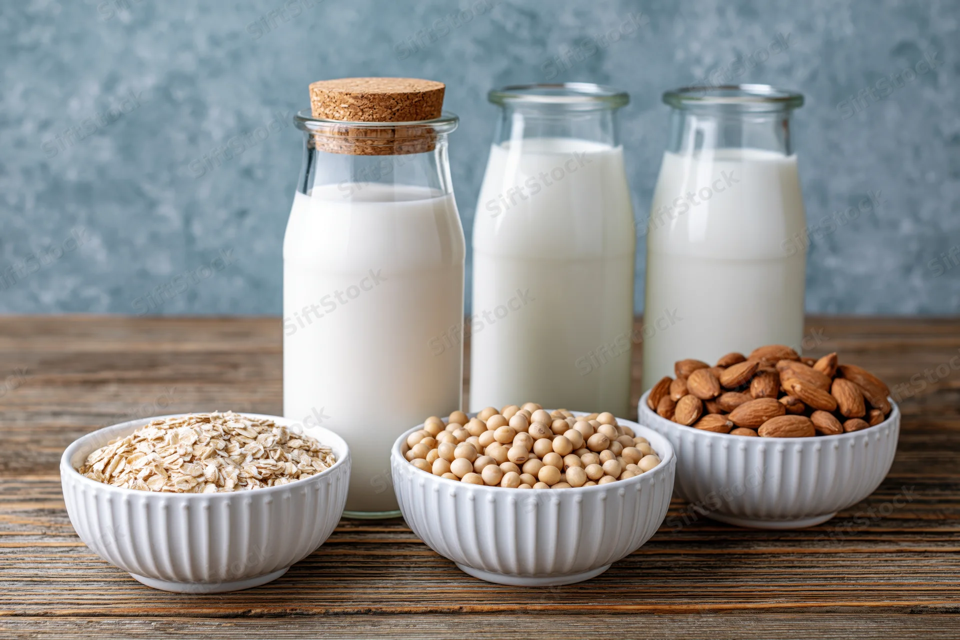 Three glass bottles of plant-based milk with bowls of oats, soybeans, and almonds on a wooden table