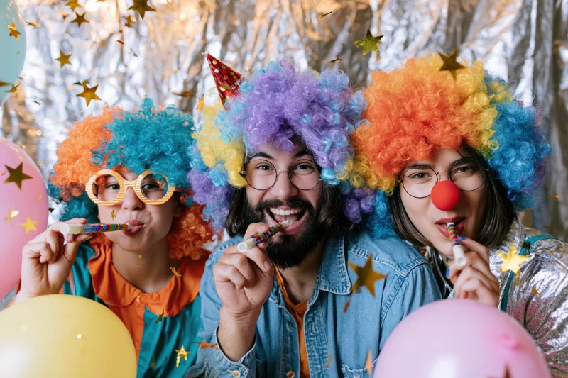Three young adults in colorful clown wigs blowing party horns at a festive celebration with balloons and confetti