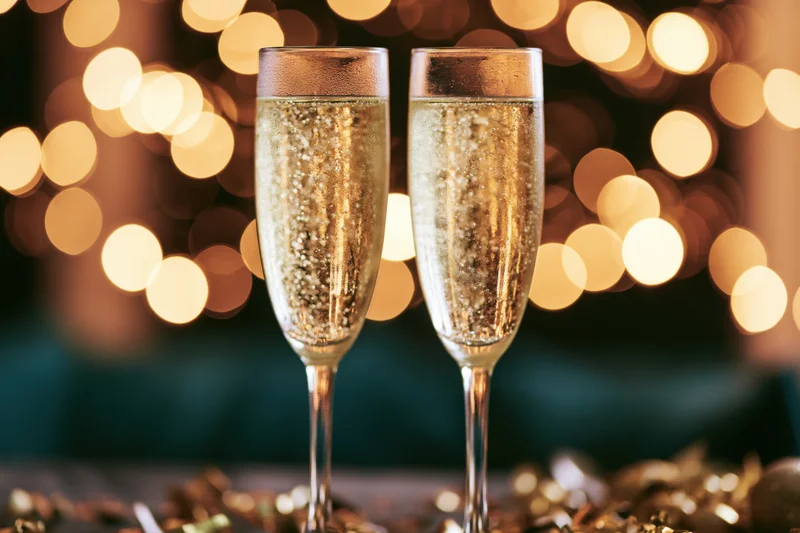 Two champagne flutes filled with sparkling wine bubbles rising, set against warm bokeh lights with golden confetti on the table
