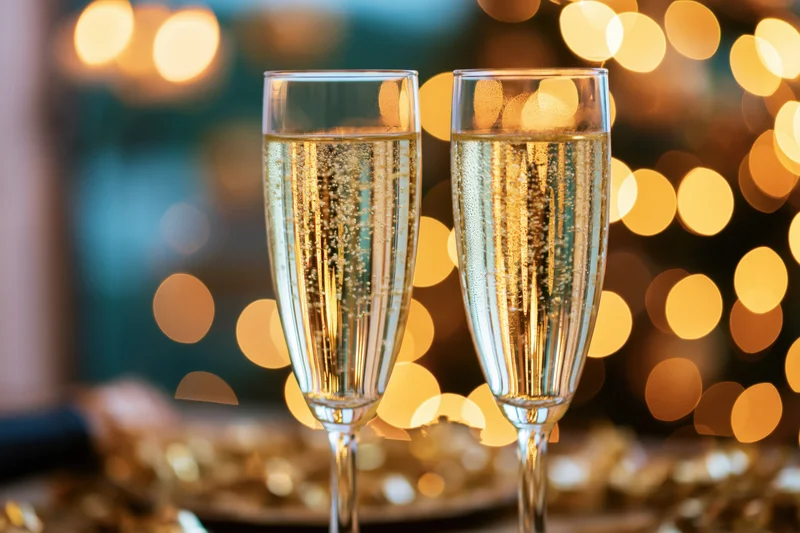 Two champagne flutes filled with sparkling wine bubbles rising, set against warm bokeh lights on a festive table