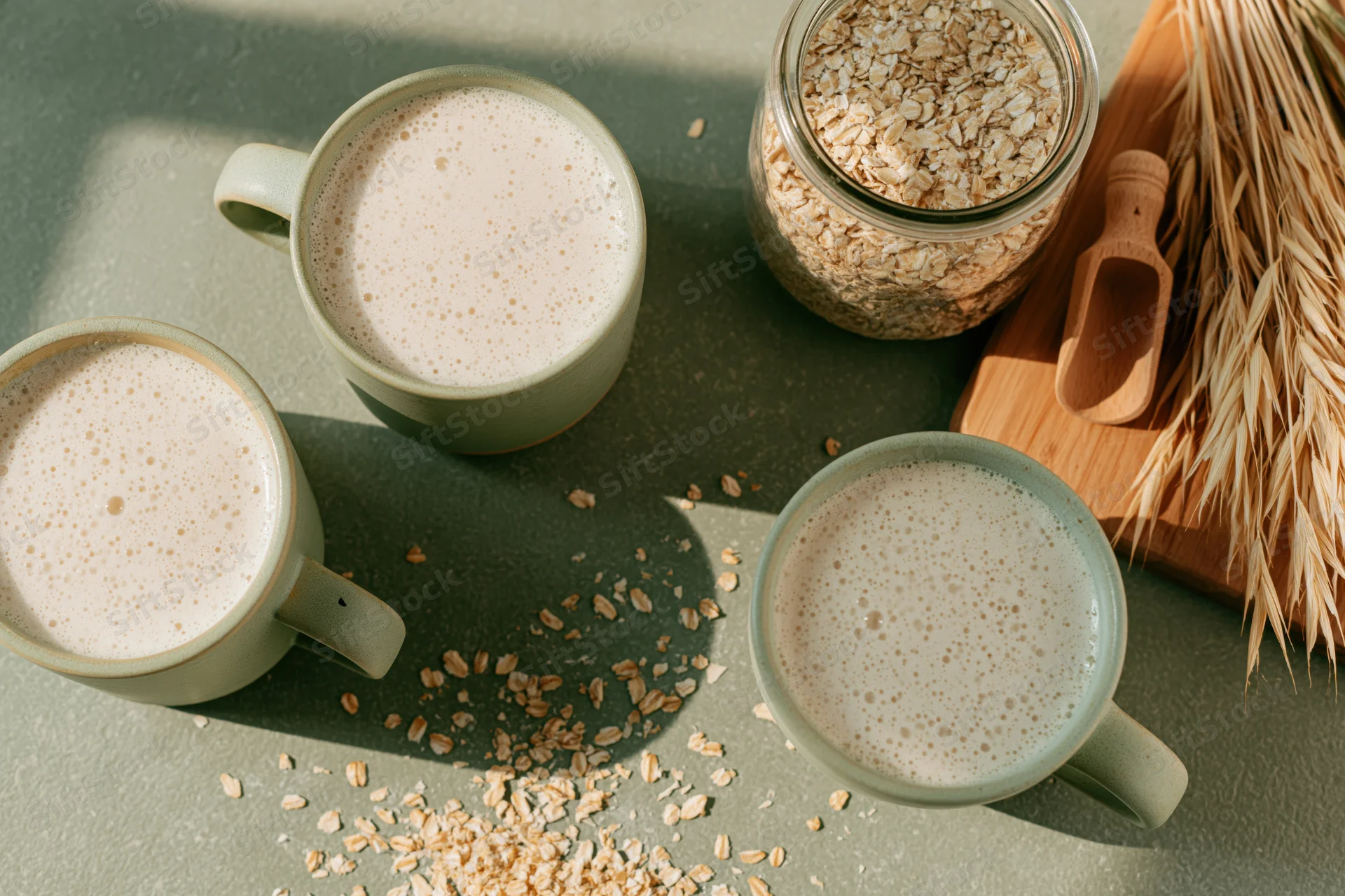 Three mugs of frothy oat milk on a green surface with scattered oats and a jar of oats nearby