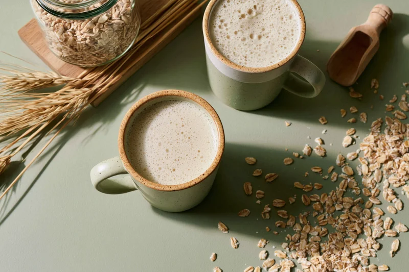 Two ceramic mugs filled with frothy oat milk on a sage-green surface with scattered oats, a glass jar, wheat stalks and a wooden scoop
