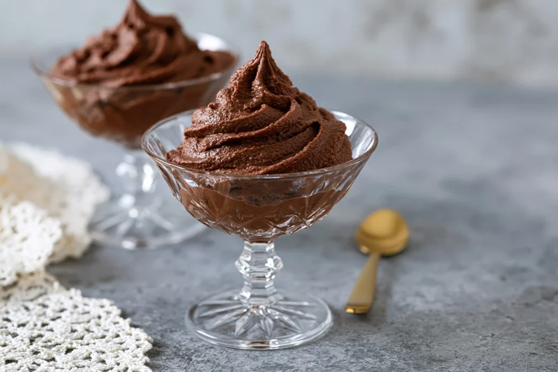 Piped chocolate mousse in glass dessert coupes on a gray countertop with a golden spoon and white lace doily