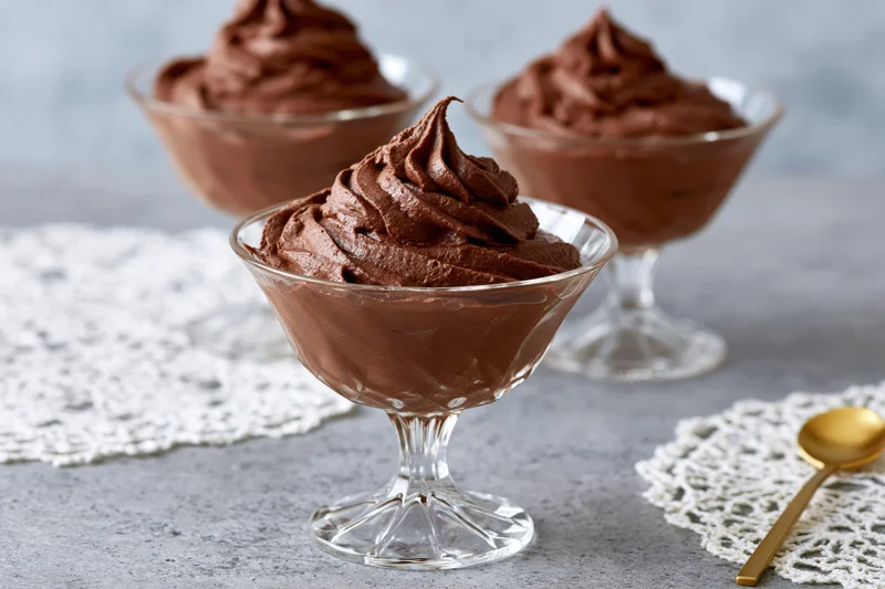 Piped chocolate mousse in glass dessert coupes on a gray surface with lace doilies and a golden spoon