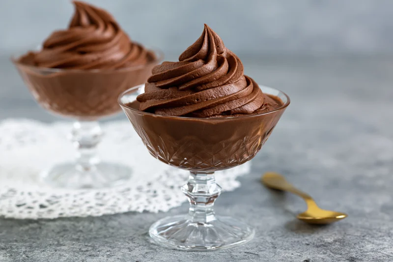 Two glass dessert coupes filled with piped chocolate mousse on a gray surface with a white lace doily and golden spoon beside