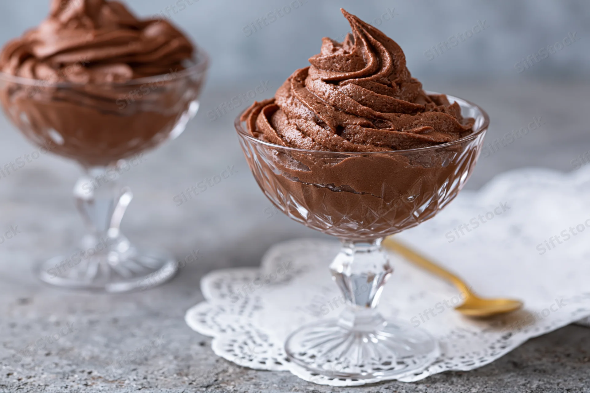 Piped chocolate mousse in glass dessert coupes on a gray table with a white doily and gold spoon