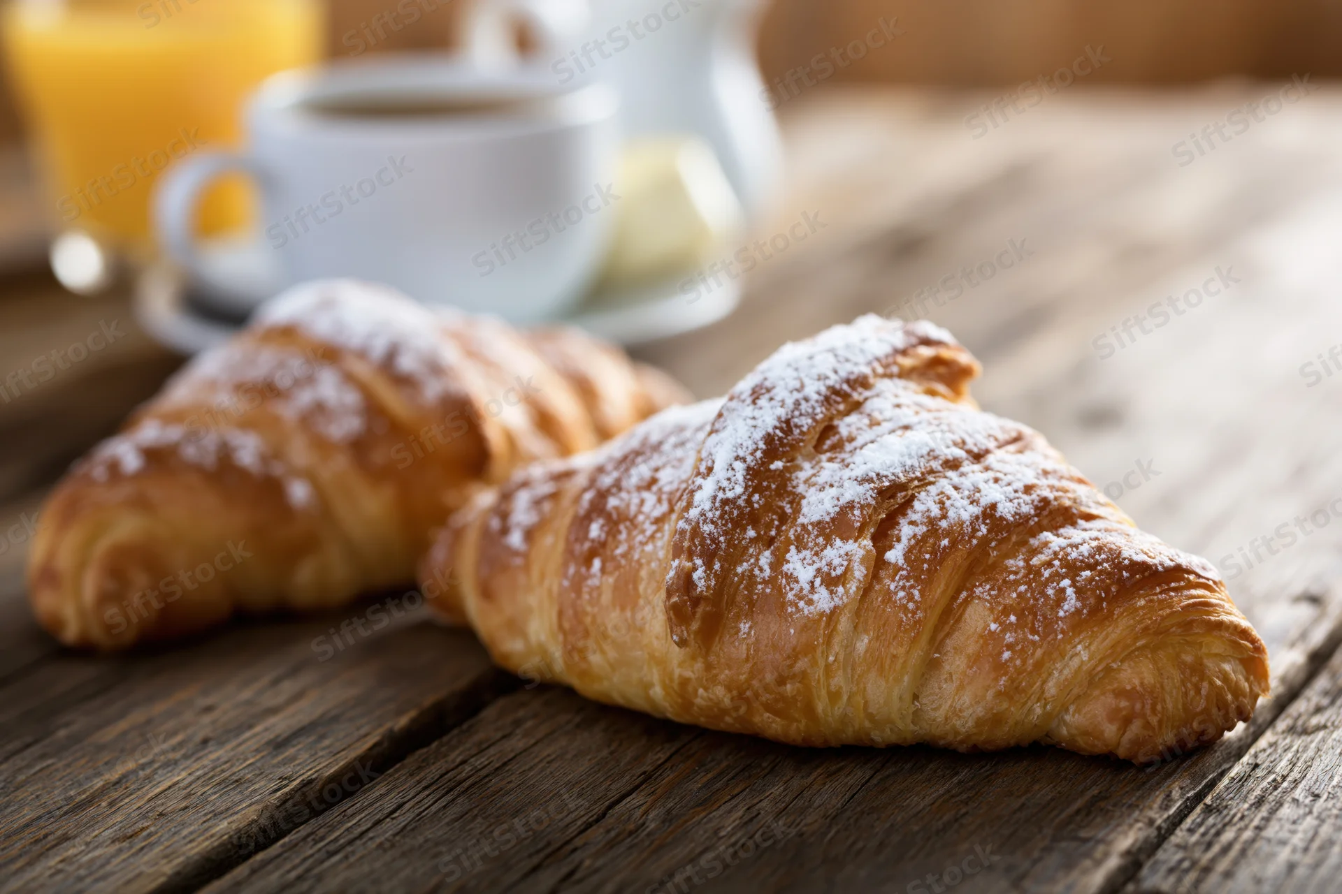 Two golden-brown croissants dusted with powdered sugar on a wooden table with coffee and orange juice in the background