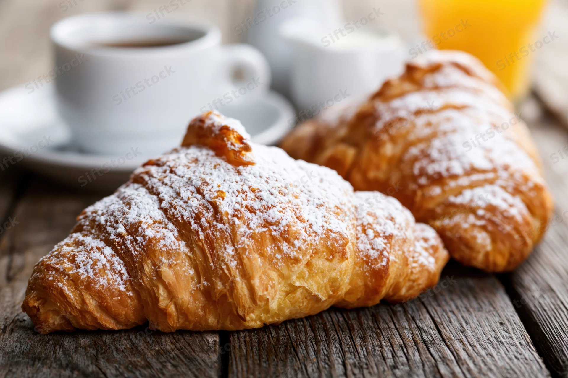 Two golden-brown croissants dusted with powdered sugar on a wooden table with coffee and orange juice in the background