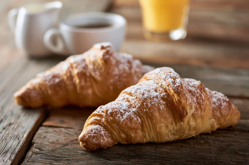Two golden-brown croissants dusted with powdered sugar on a rustic wooden table with coffee and orange juice in the background
