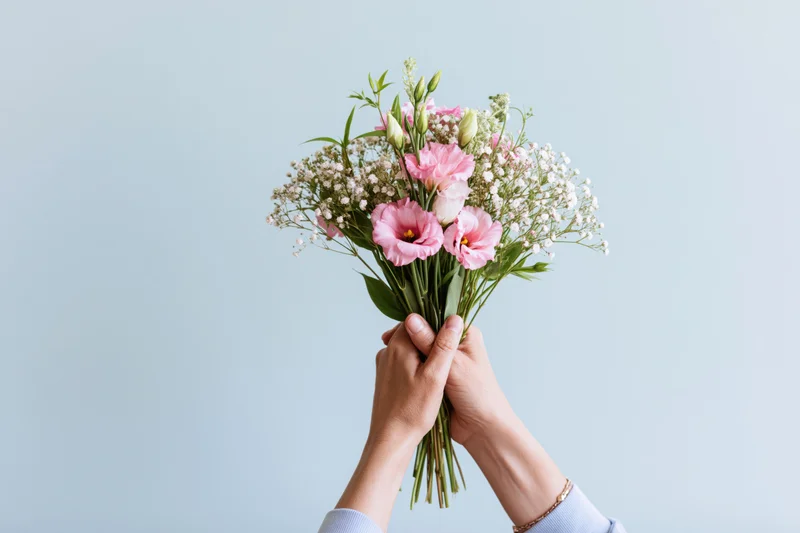 Hands holding a bouquet of pink eustoma and white baby's breath against a light blue background