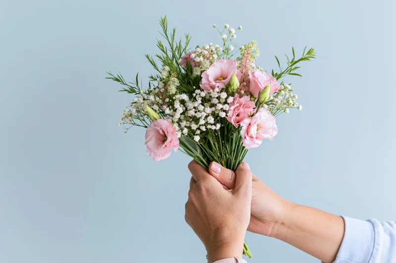 Hands holding a bouquet of pink eustoma and white baby's breath against a light blue background