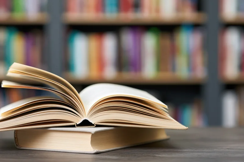 Open hardcover book resting on another book on a dark wooden table with bookshelves in the background