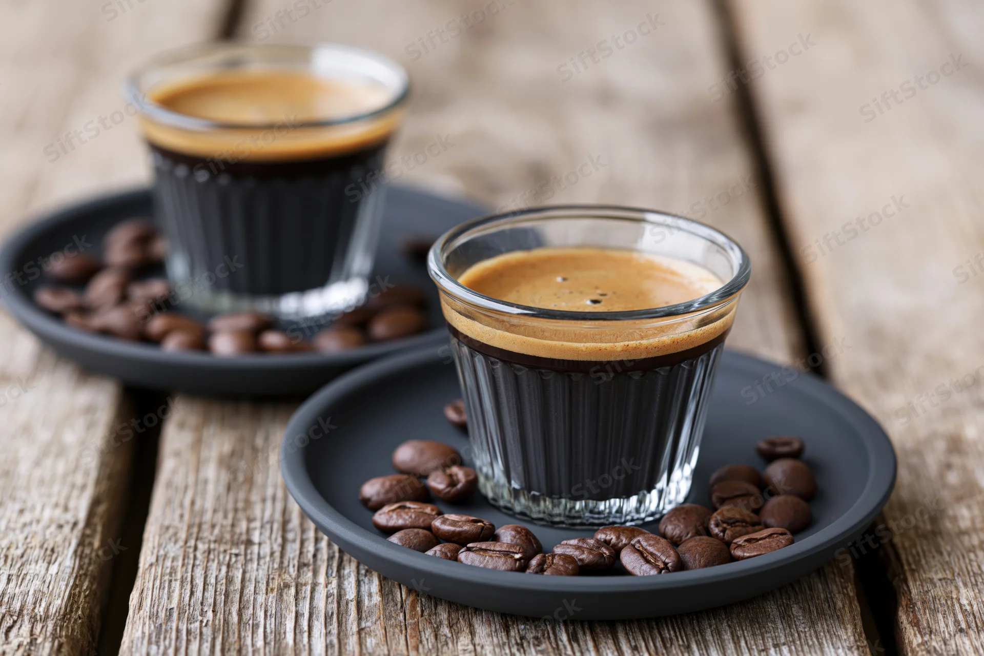 Two glass espresso cups filled with dark coffee and crema on black saucers with roasted beans on a wooden table