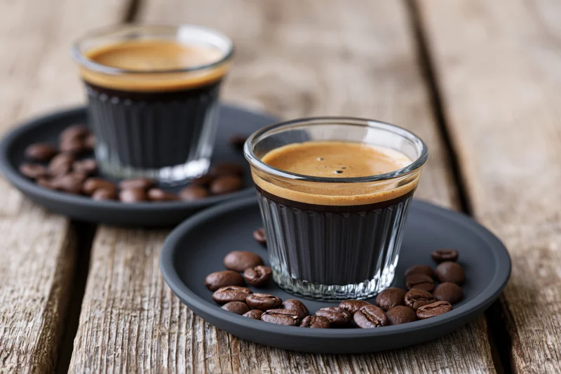 Two glass espresso cups filled with dark coffee and crema on black saucers with roasted beans on a wooden table