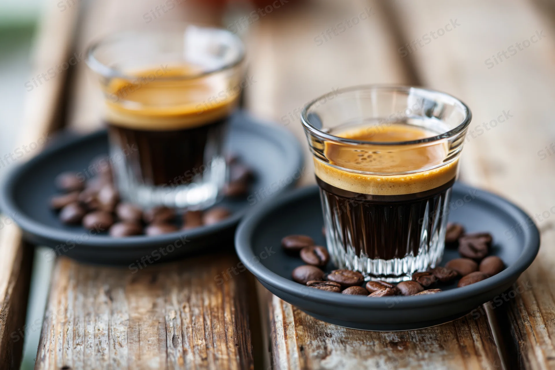 Two espresso glasses with crema on black saucers surrounded by coffee beans on a wooden tray