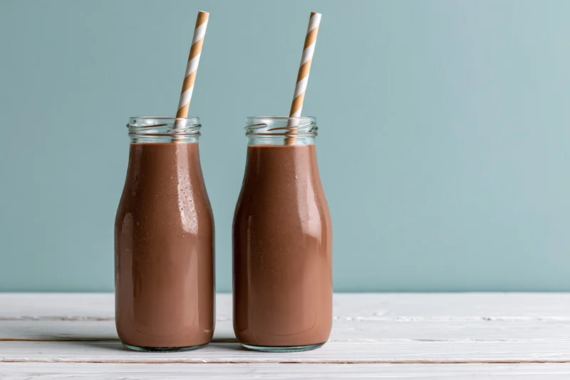 Two glass bottles of chocolate milk with striped straws on a white wooden table