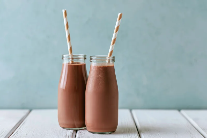 Two glass bottles filled with chocolate milk on a white wooden table with striped paper straws