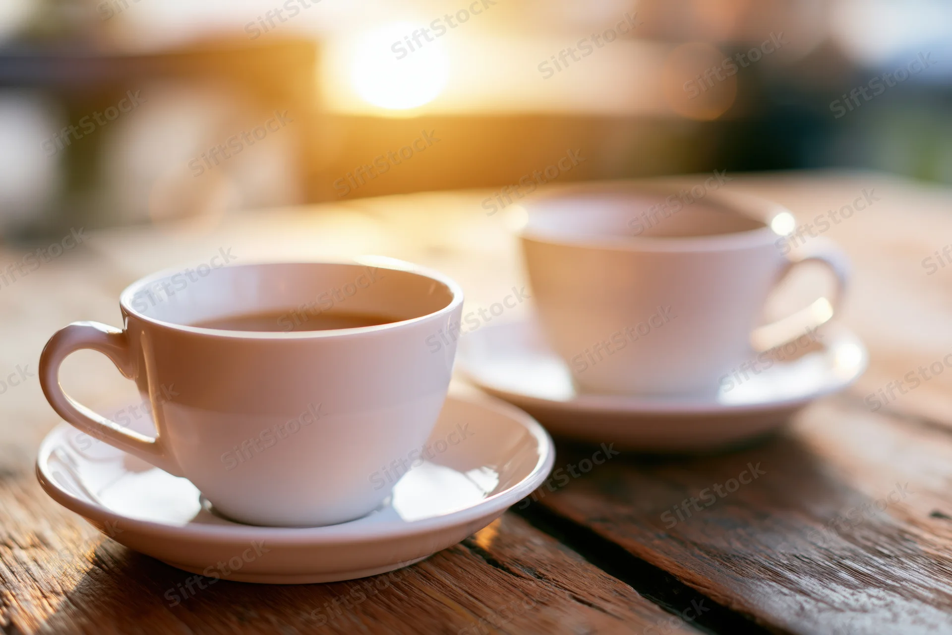 Two white ceramic coffee cups on saucers resting on a worn wooden table in warm morning light