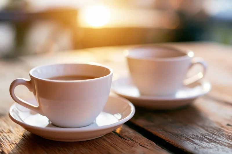 Two white ceramic coffee cups on saucers resting on a worn wooden table in warm morning light