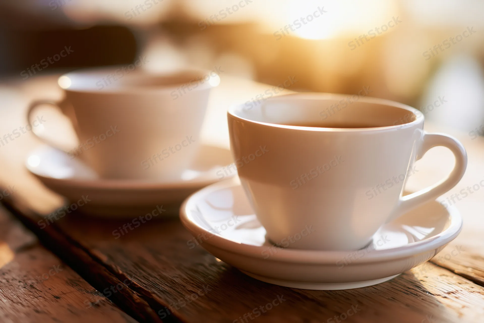 Two white ceramic coffee cups on saucers resting on a worn wooden table in warm morning light