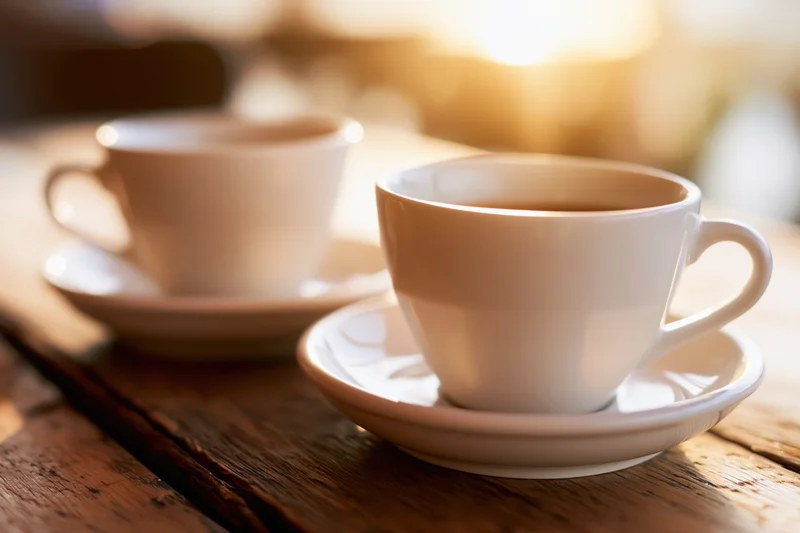 Two white ceramic coffee cups on saucers resting on a worn wooden table in warm morning light