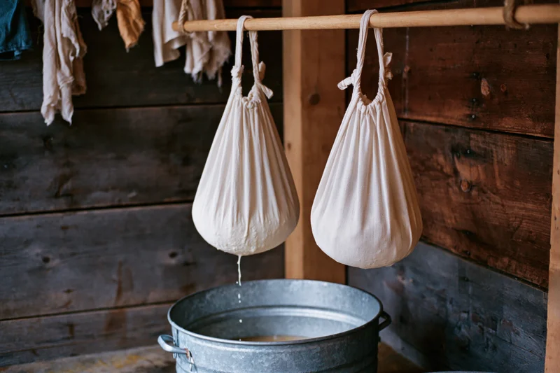 Two white cloth bags filled with curds hanging over a metal bucket in a rustic wooden farmhouse kitchen