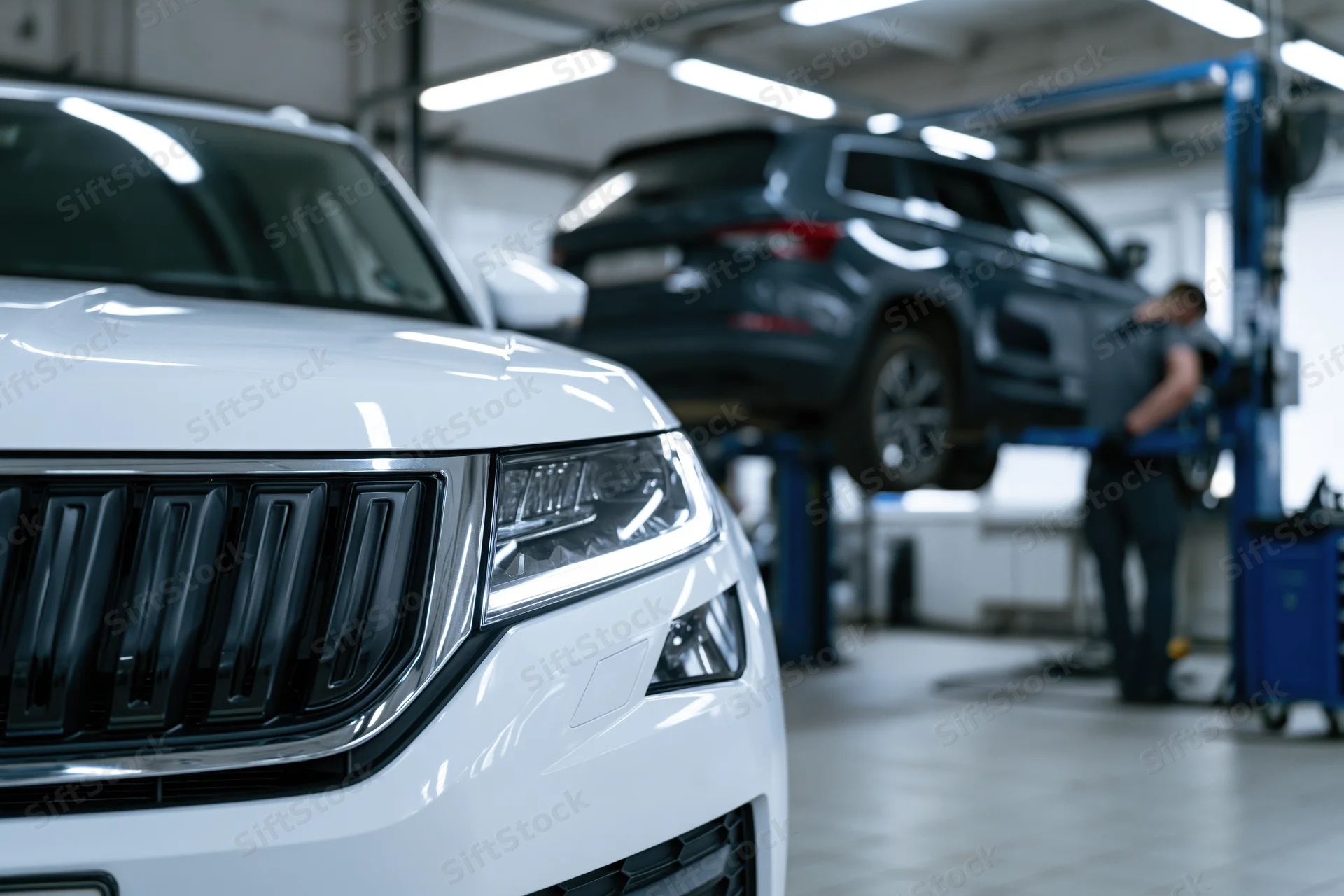 White suv front end in focus with mechanic working on blue suv on lift in background garage