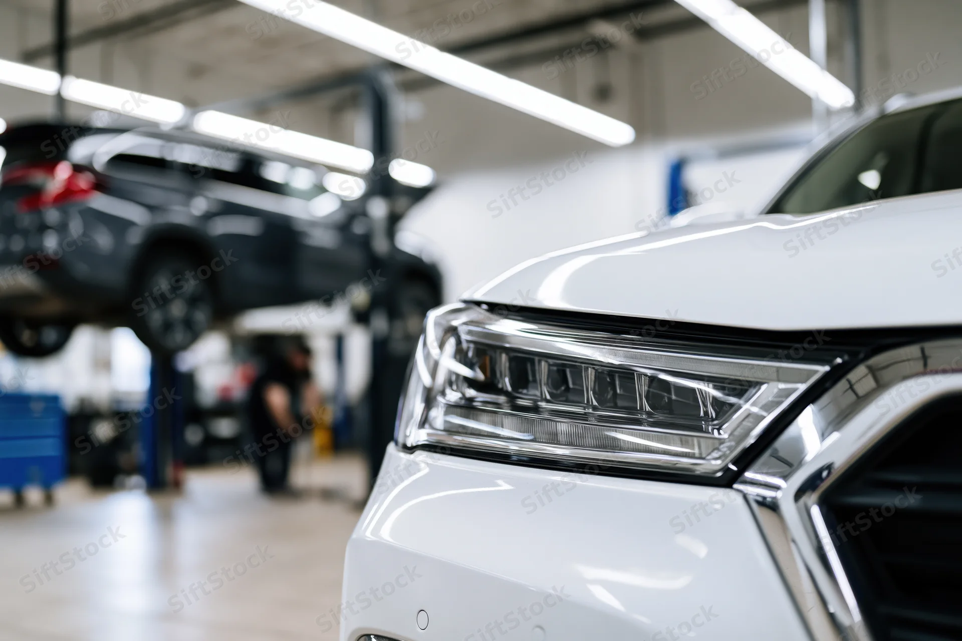 White suv front end in focus with mechanic working on elevated vehicle in background garage