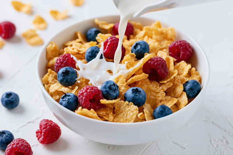 Milk being poured into a bowl of cornflakes with blueberries and raspberries on a white surface