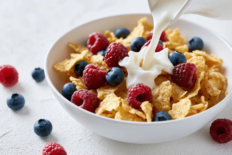 Milk being poured into a bowl of cornflakes with blueberries and raspberries on a white surface
