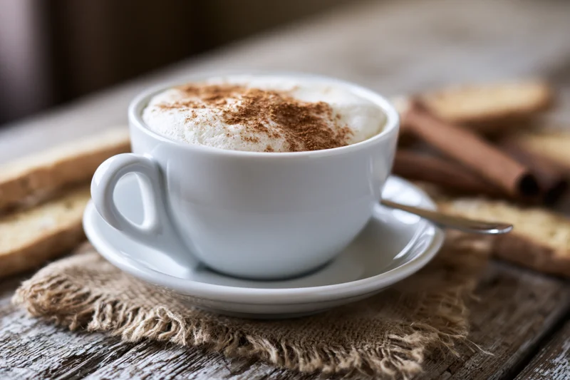 White ceramic cappuccino cup with thick milk foam and cinnamon dust on a wooden table with biscotti and cinnamon sticks