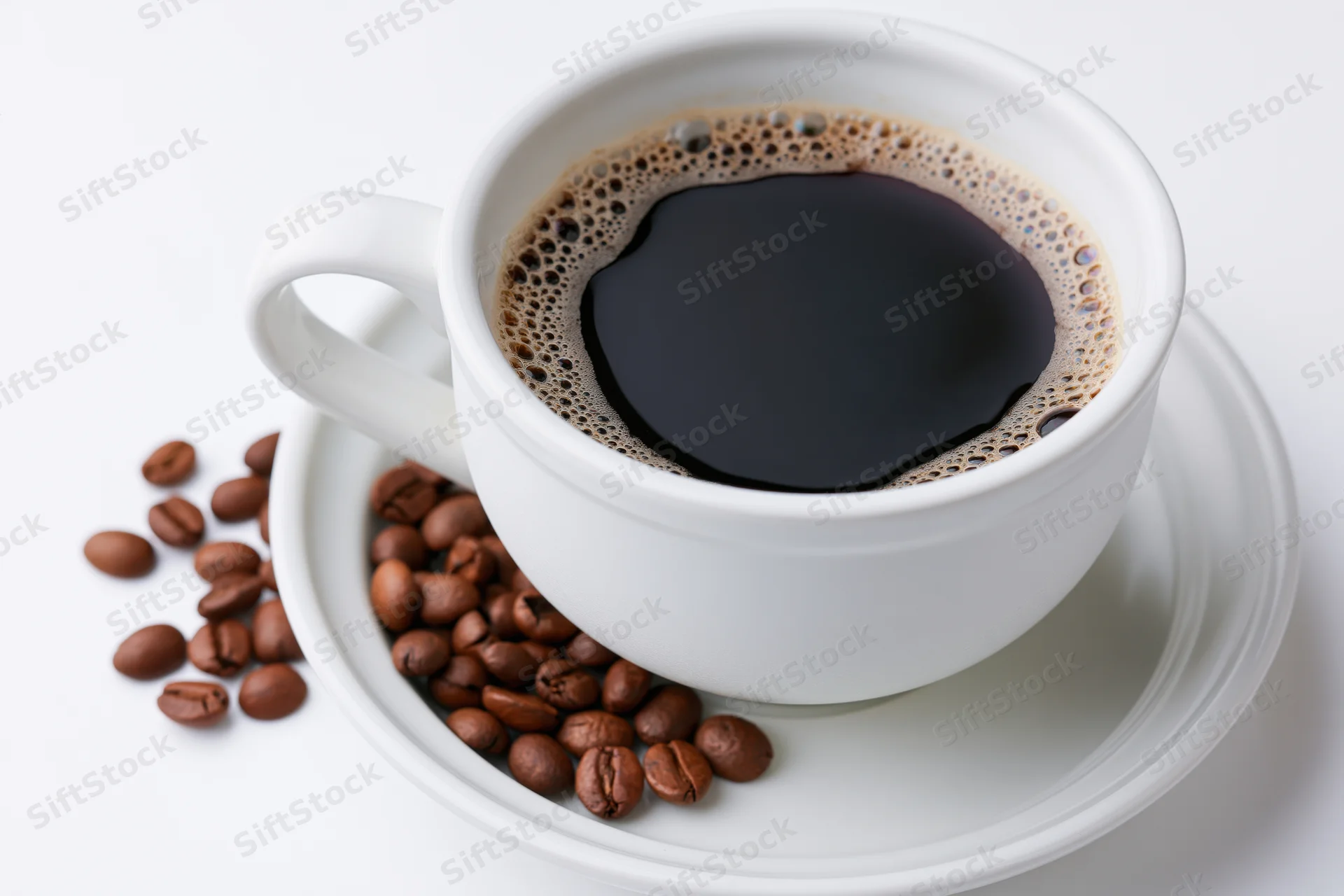 White ceramic coffee cup filled with black coffee on matching saucer with scattered beans around it