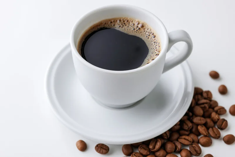 White ceramic coffee cup filled with black coffee on matching saucer with scattered roasted beans on white surface