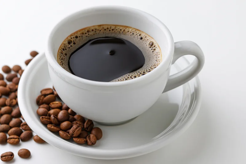 White ceramic coffee cup filled with black coffee on matching saucer with scattered beans on white surface