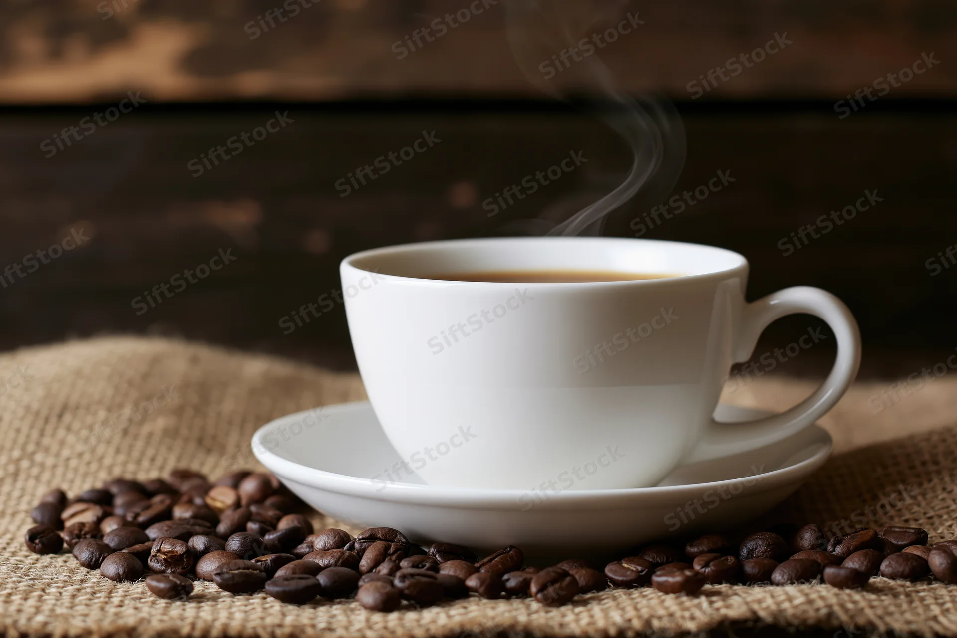 Steaming white ceramic coffee cup on saucer surrounded by roasted beans on burlap cloth