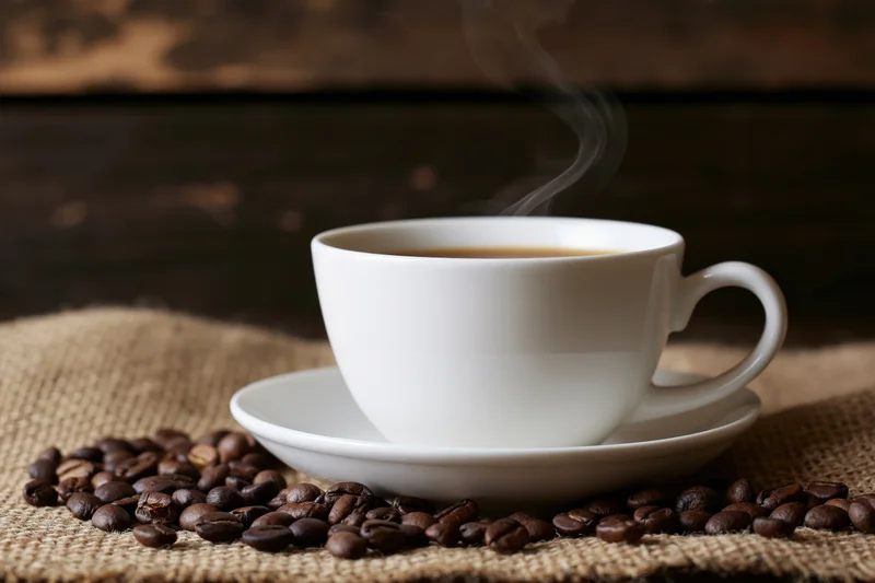 Steaming white ceramic coffee cup on saucer surrounded by roasted beans on burlap cloth