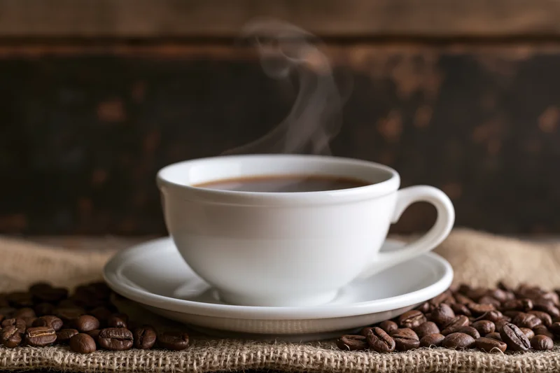 Steaming white ceramic coffee cup on saucer surrounded by roasted beans on burlap cloth