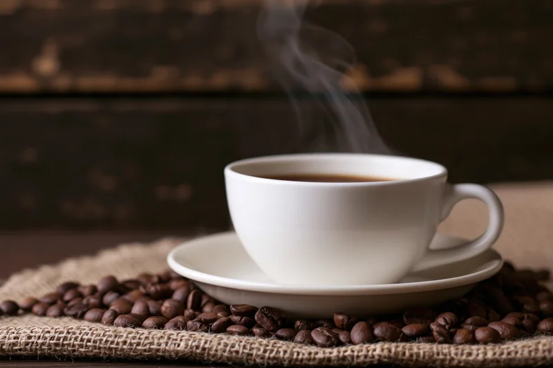 Steaming white ceramic coffee cup on saucer surrounded by coffee beans on burlap cloth