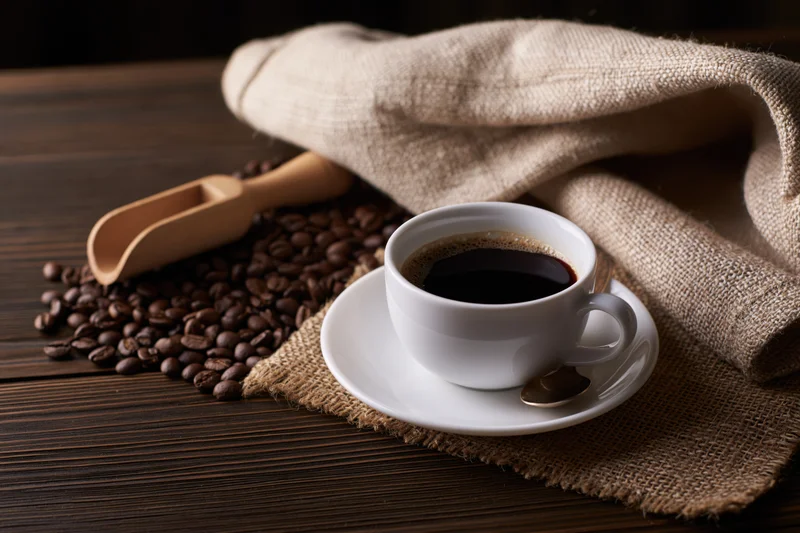 White ceramic coffee cup on saucer with coffee beans and burlap sack on wooden table