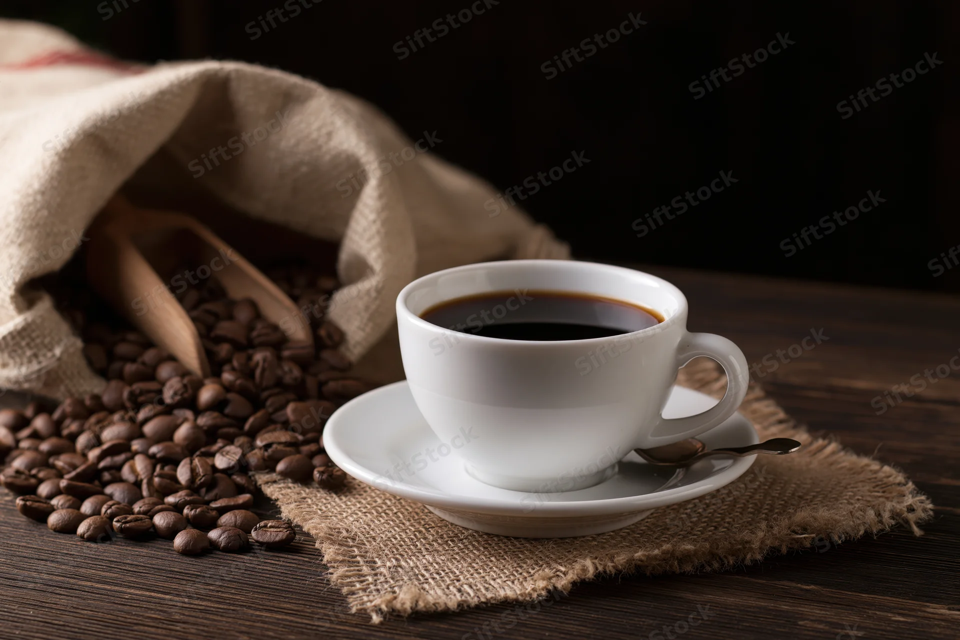 White ceramic coffee cup on saucer with coffee beans spilling from burlap bag on wooden table
